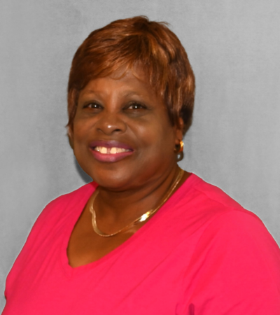 a person with a warm smile wearing short auburn hair, gold hoop earrings, a gold necklace, and a bright pink v-neck top, photographed against a neutral gray background.