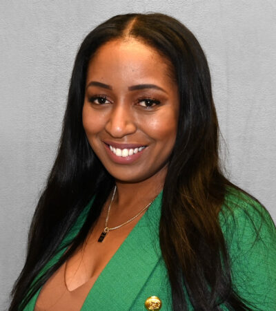 person with long straight black hair and a bright smile, wearing a green blazer with gold buttons and a delicate gold necklace, photographed against a neutral gray background.